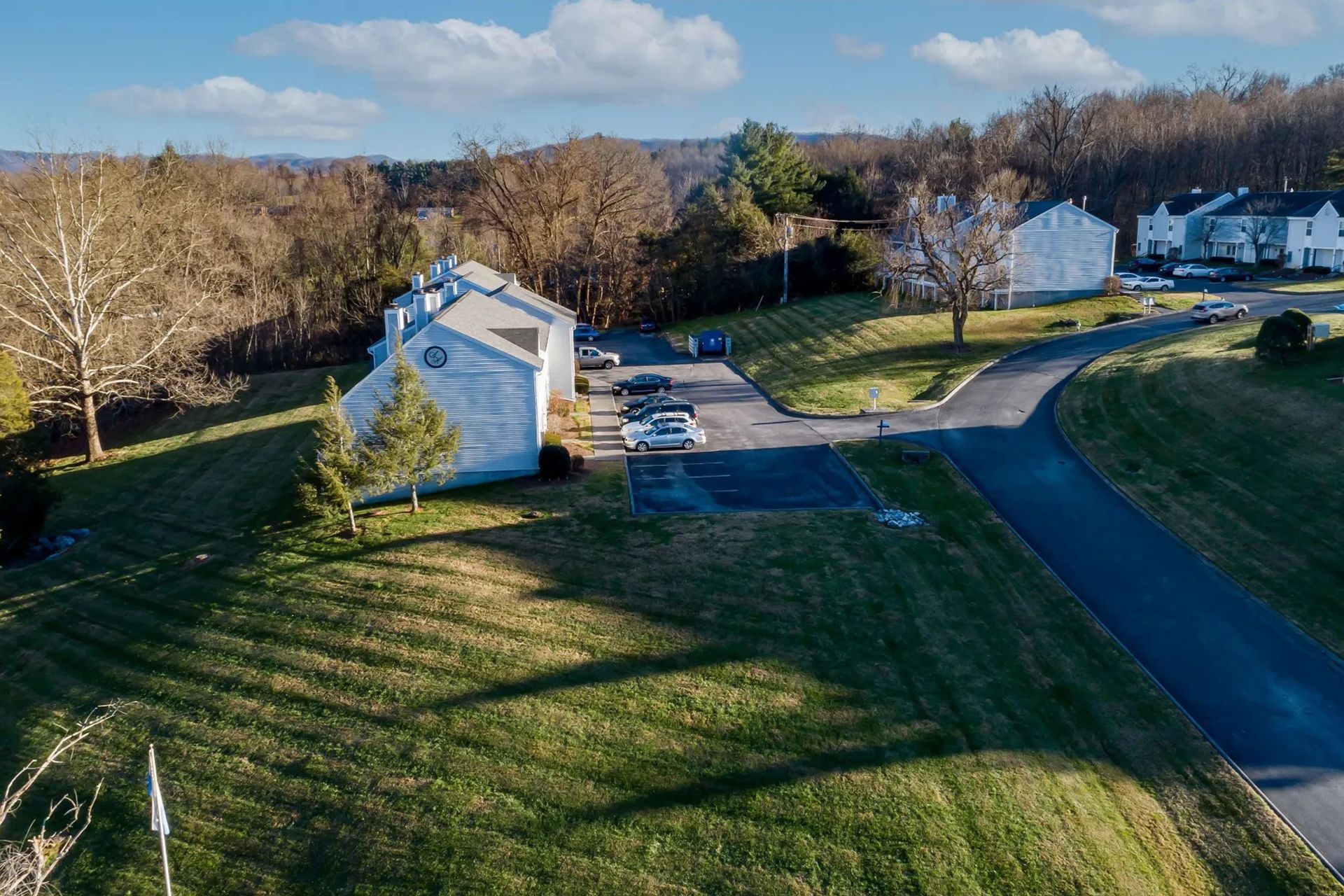 Residential buildings with cars in parking spots and a winding road, surrounded by trees and green grass under a cloudy sky.