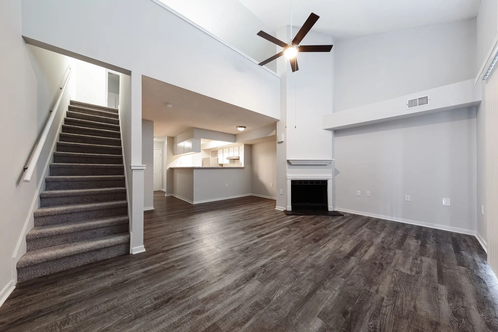 Spacious living room with a staircase, kitchen, and a fireplace. Wooden floors, white walls, and a ceiling fan.