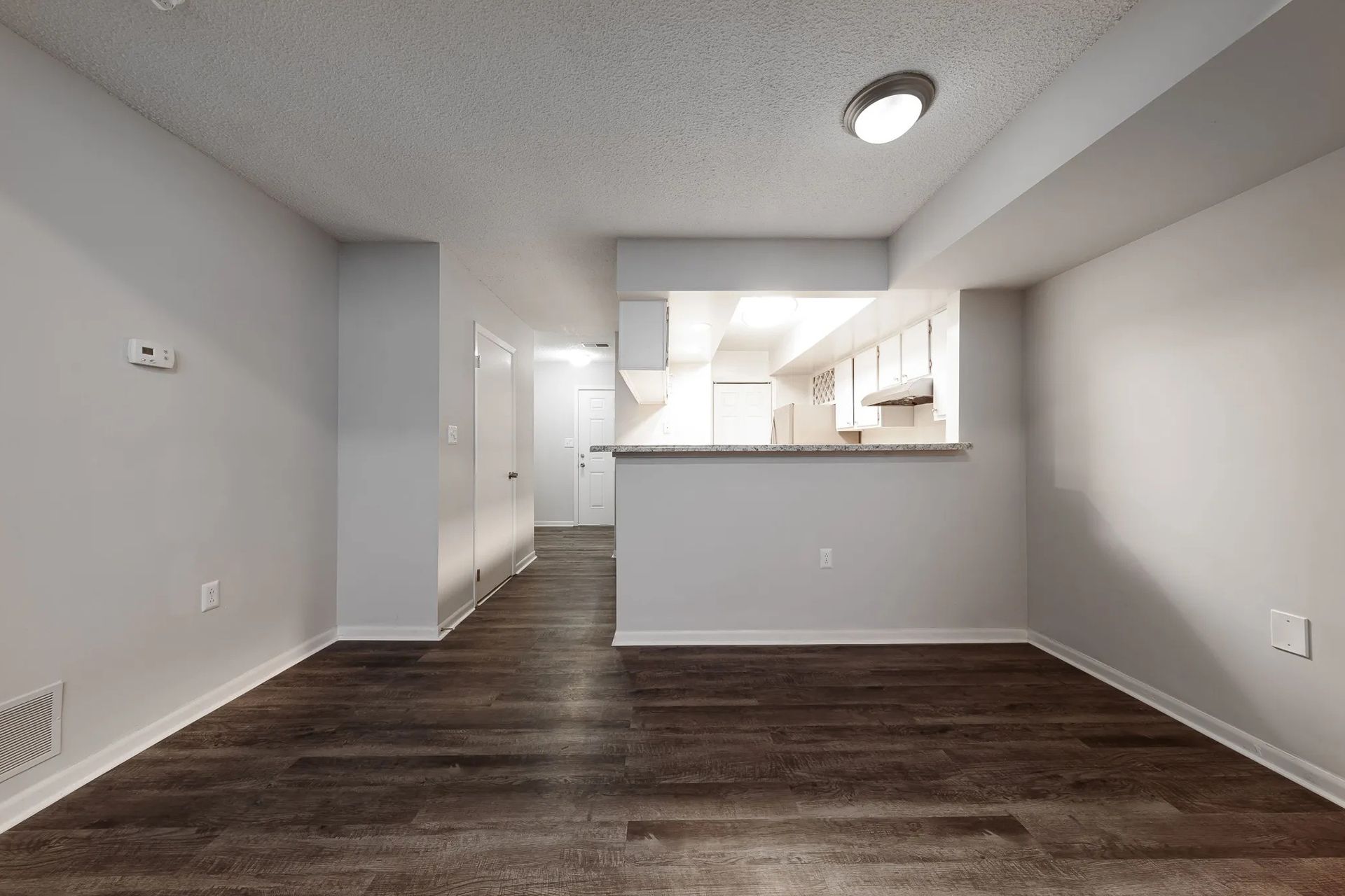 Empty apartment interior with wood-look flooring, light gray walls, and a view into the kitchen.