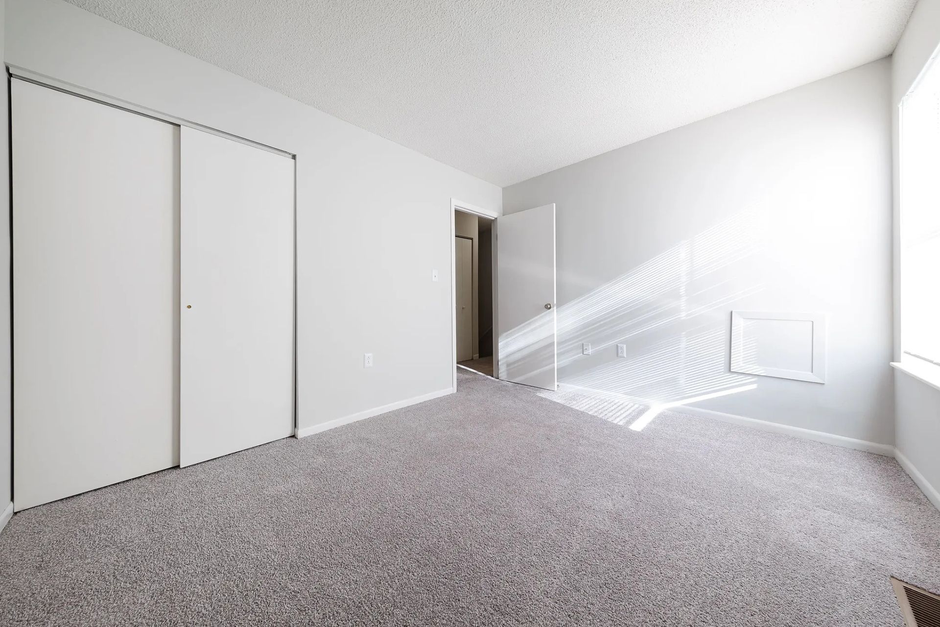 Empty bedroom with gray walls, carpet, closet doors, and a bright window.