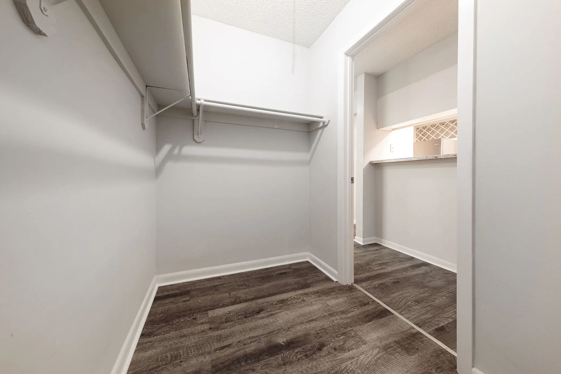 Empty walk-in closet with wood-look flooring and open doorway to a small room, white walls.