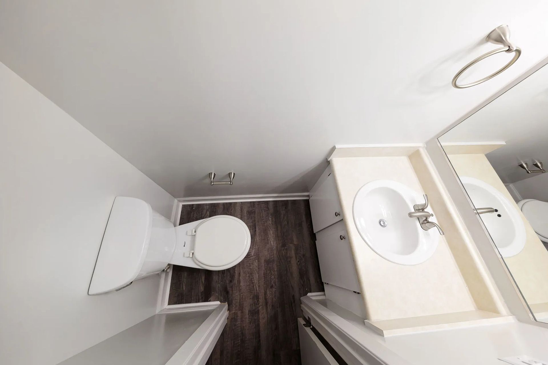 Overhead view of a small bathroom with a toilet, sink, mirror, and dark wood-look flooring.