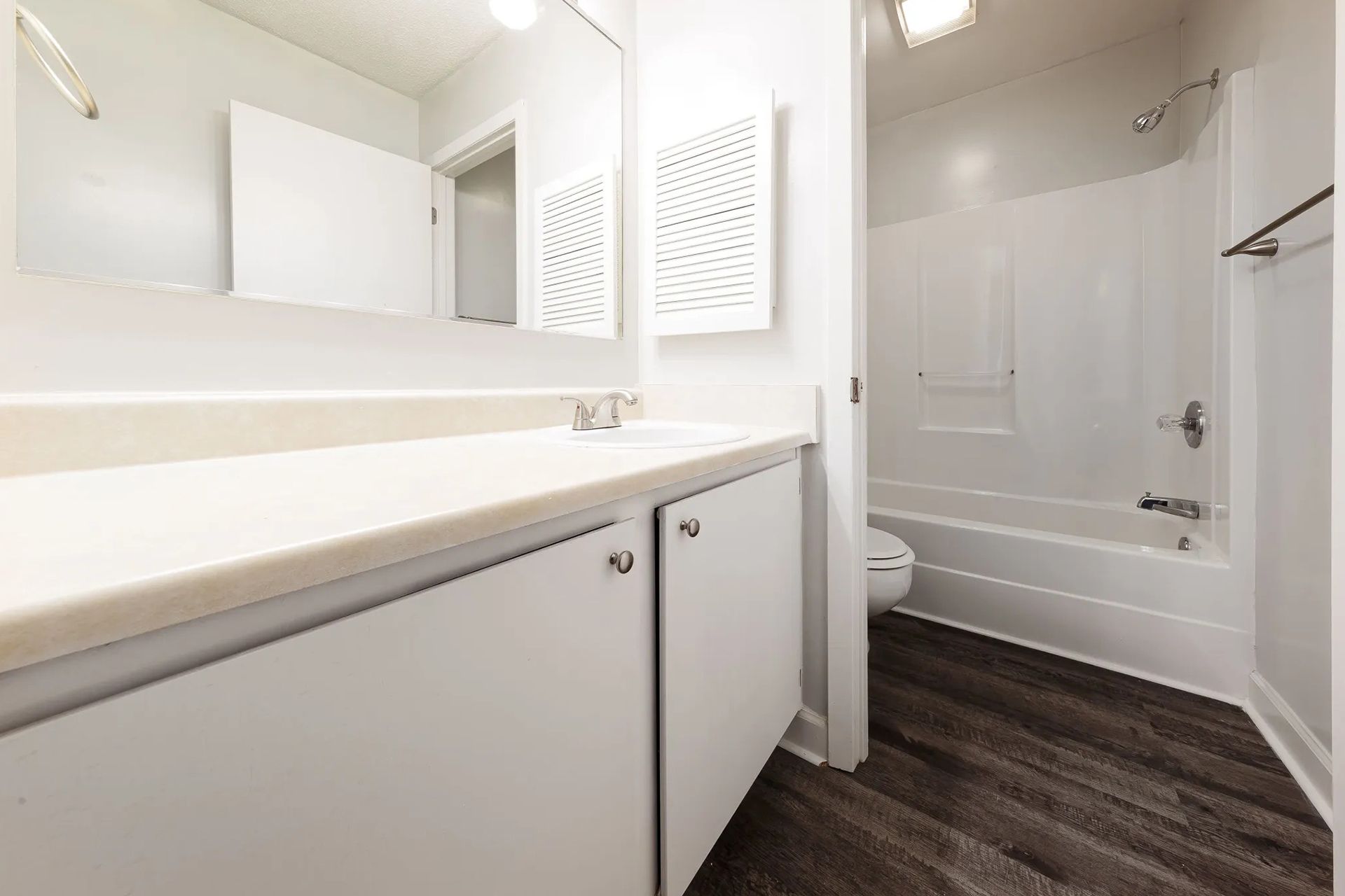 White bathroom with a vanity, toilet, and shower. Gray wood-look flooring.