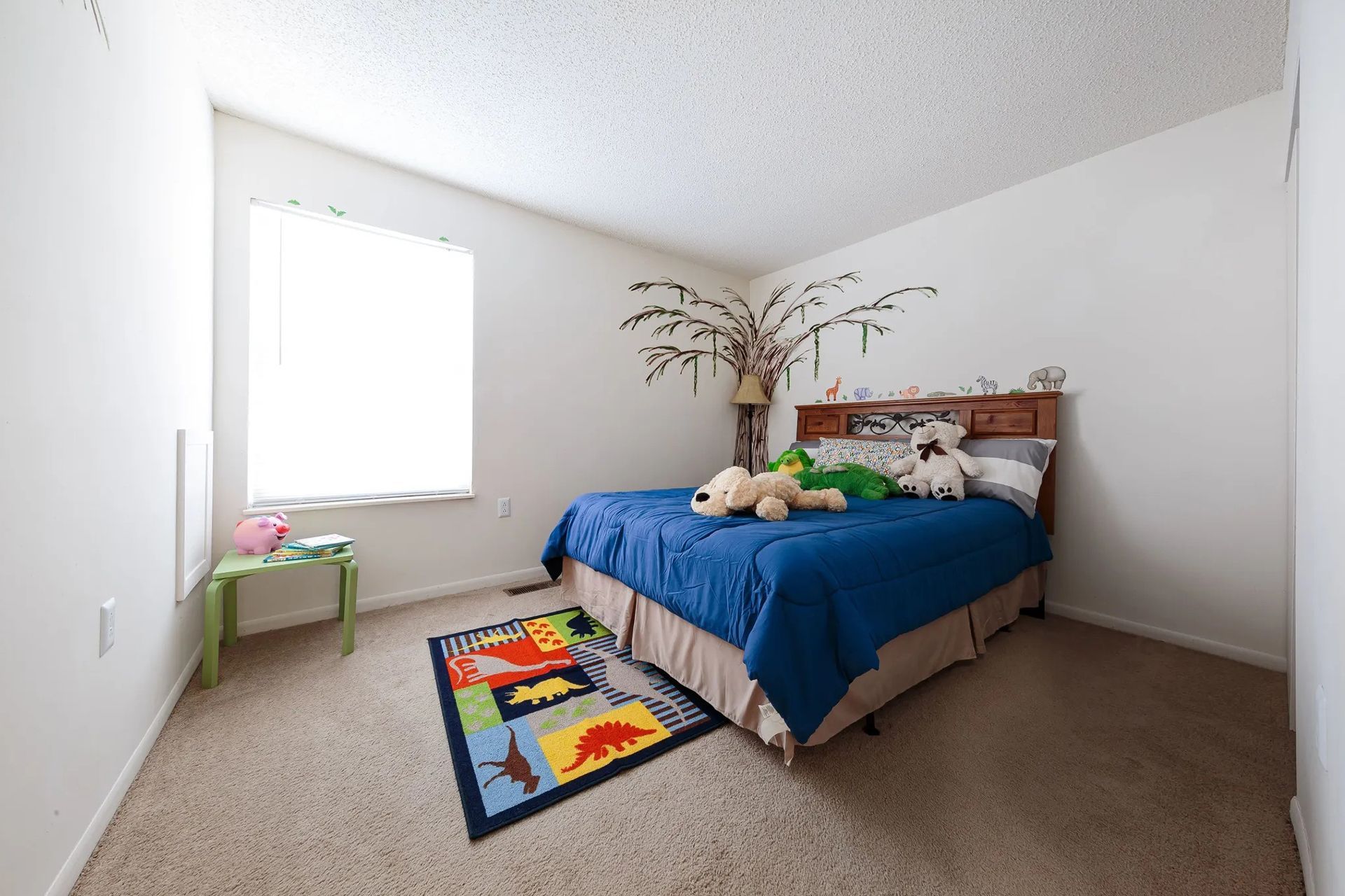 A child's bedroom with a bed, rug, and a window. Toys and a tree decal decorate the room.