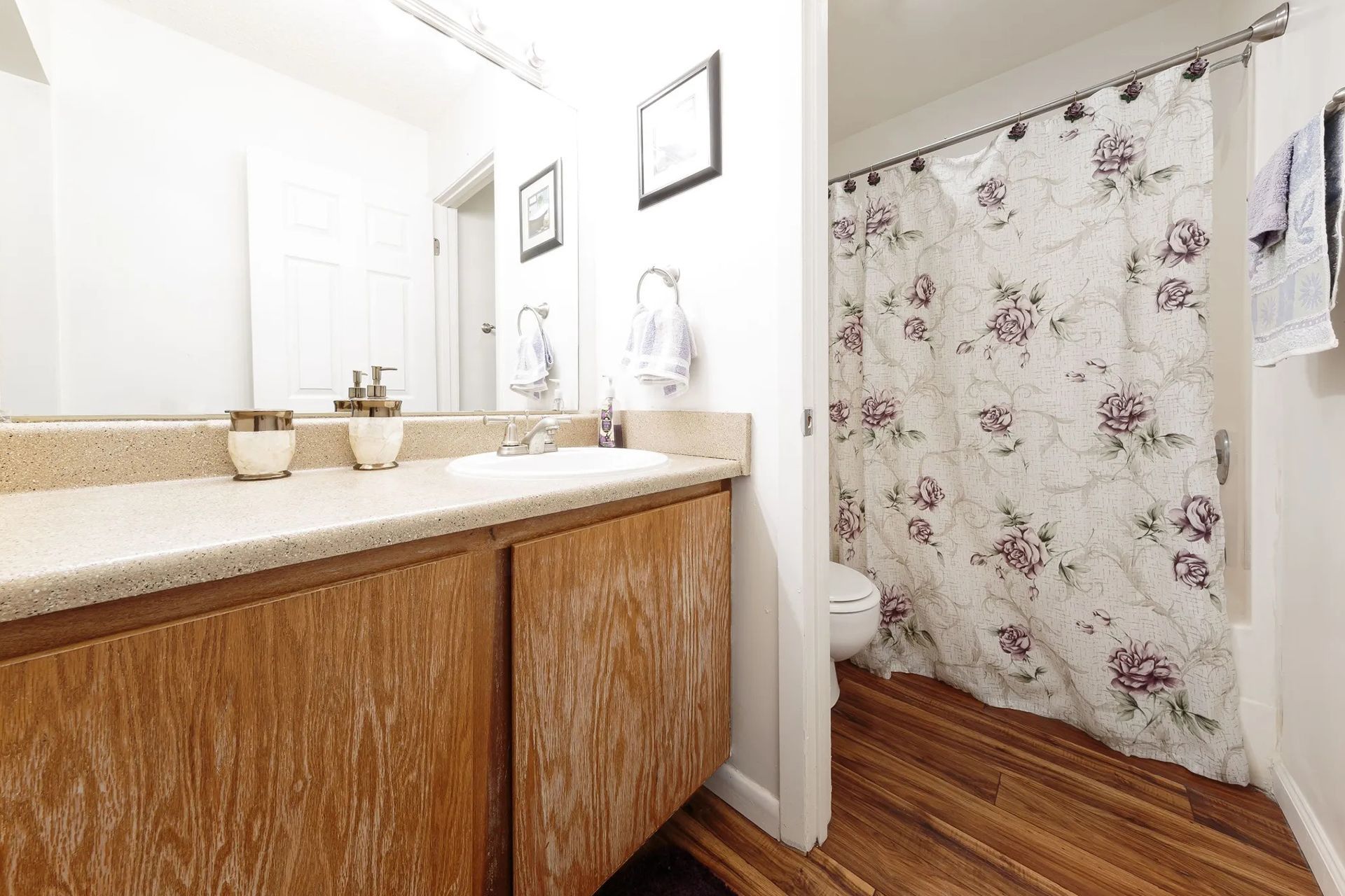 Bathroom with wooden cabinets, white walls, floral shower curtain, and wooden floor.