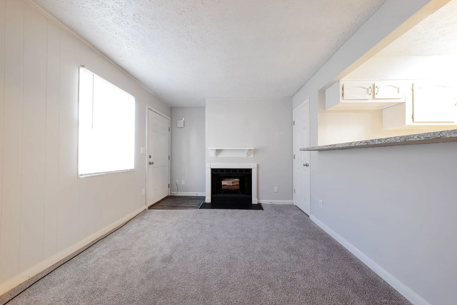 Living room with fireplace, window, and partial kitchen view. Light gray walls, carpet, and cabinets.