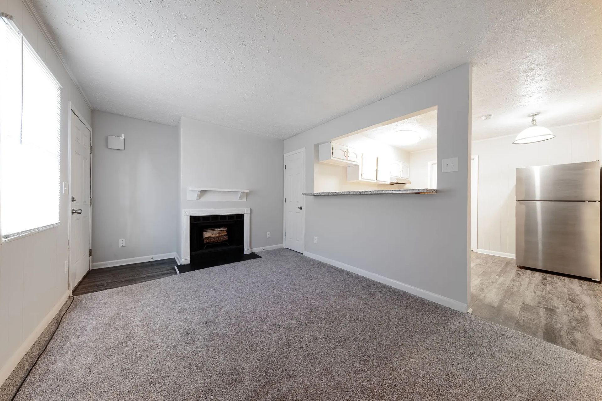 Cozy living room with fireplace, kitchen pass-through, and stainless steel refrigerator. Gray carpet and white walls.