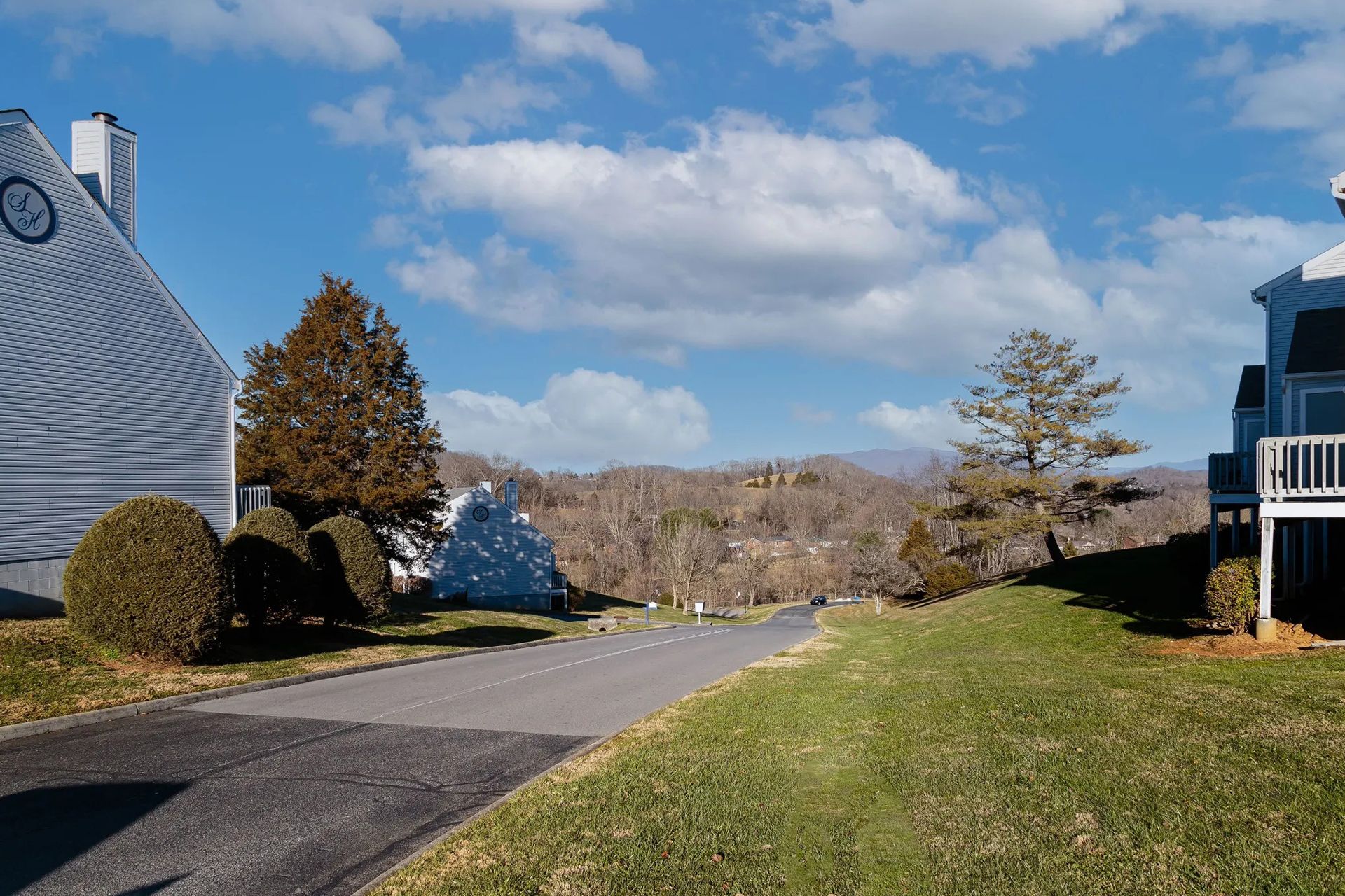 Paved road between two buildings with grass on the right, view of mountains in the distance, under a blue sky with clouds.
