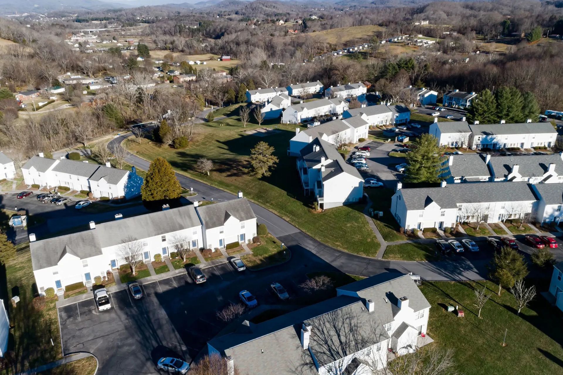 Aerial view of a residential neighborhood with white townhouses, cars, and a green grassy area. Mountains in the background.
