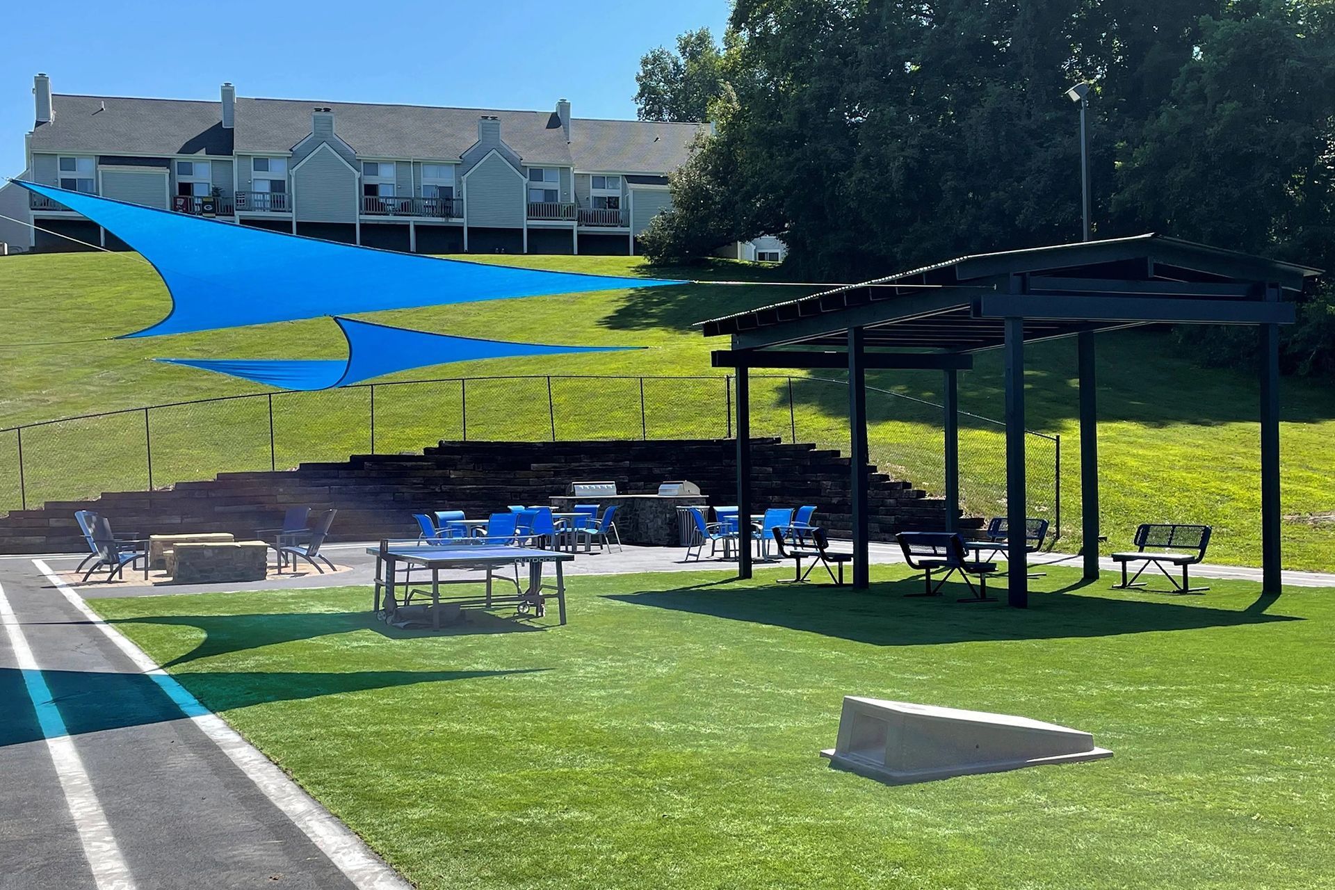 Outdoor recreation area with picnic tables, grill, gazebo, blue shade, grassy lawn, and apartments on a hill.