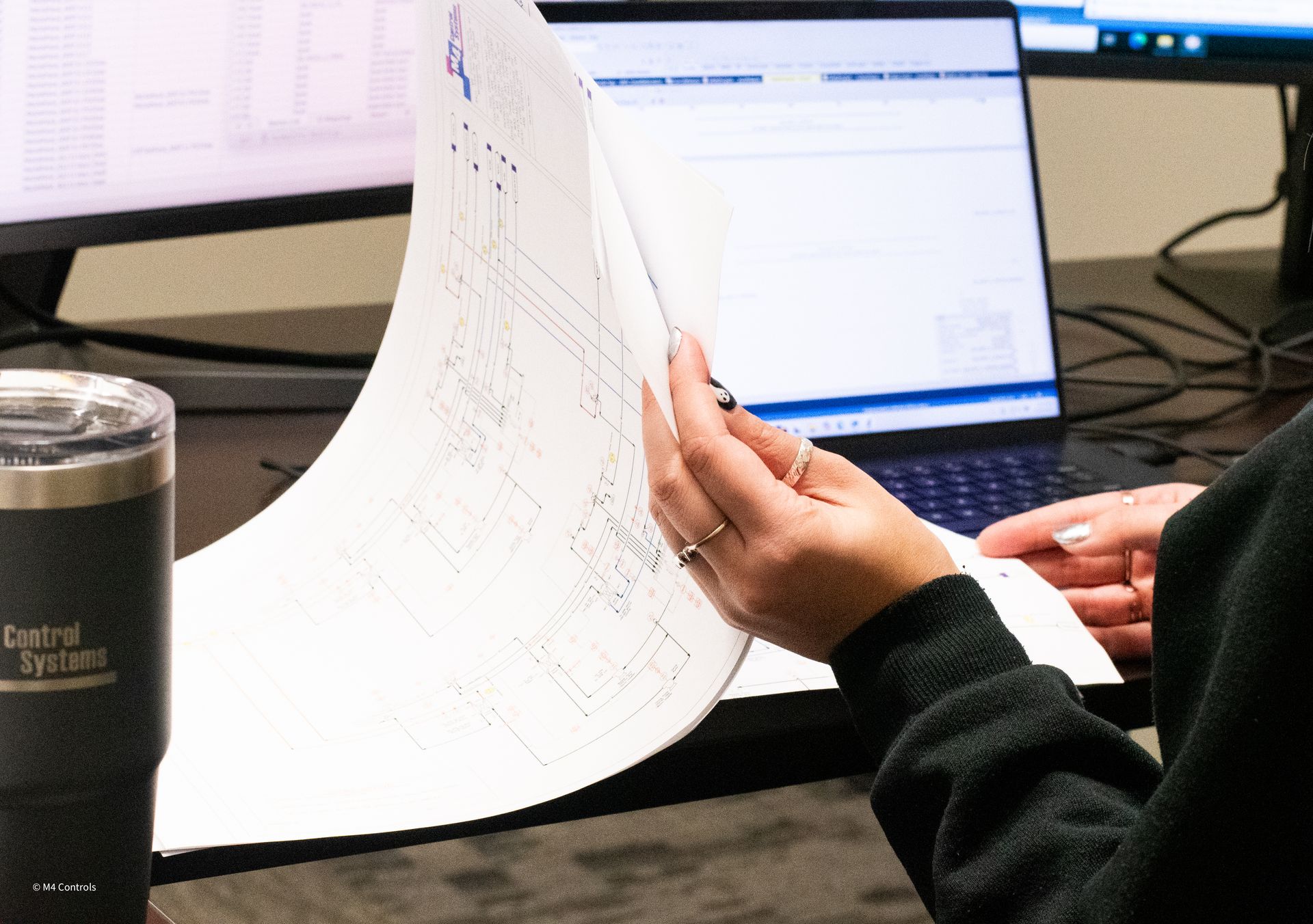 Person holding blueprints, comparing them to a computer screen in an office. A tumbler is in the foreground.