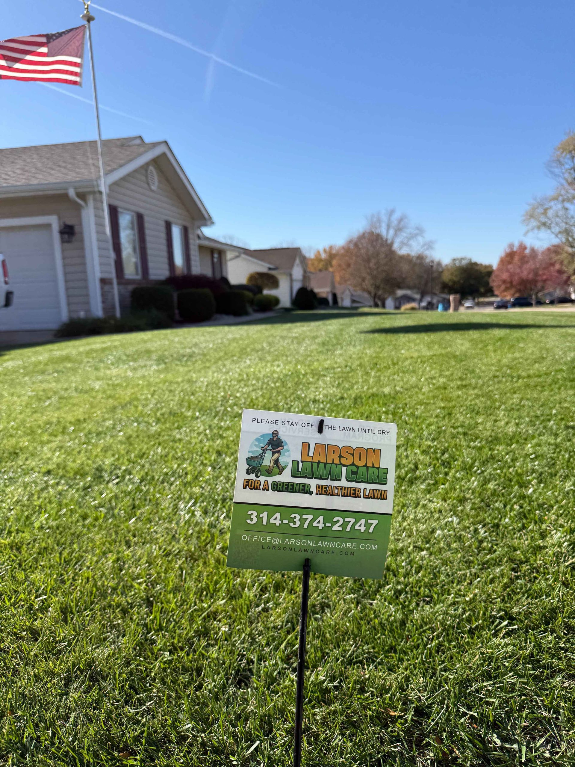 Sign in a well-manicured lawn advertising lawn care services in front of a house, American flag in the distance.