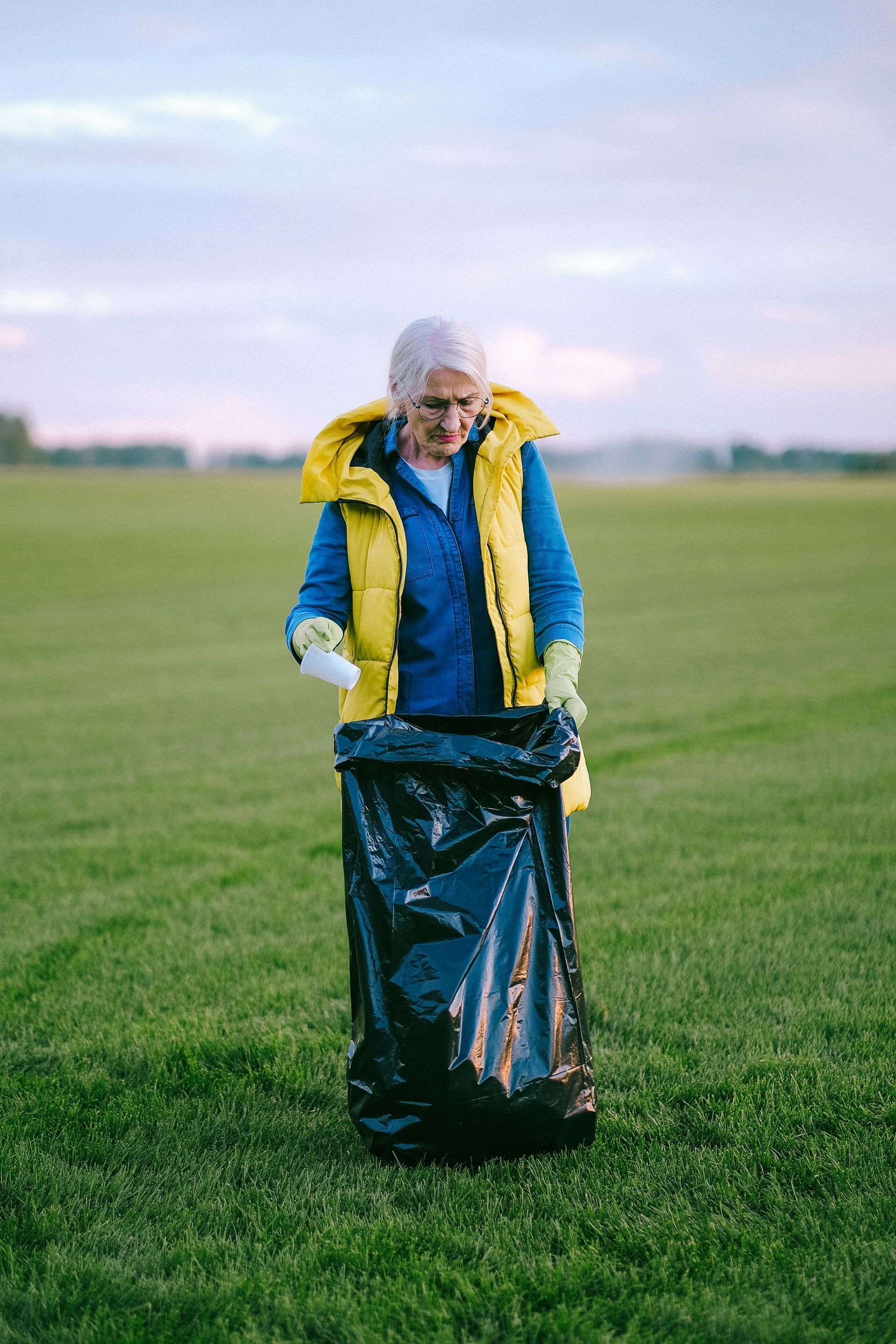 An elderly woman is picking up trash in a field.