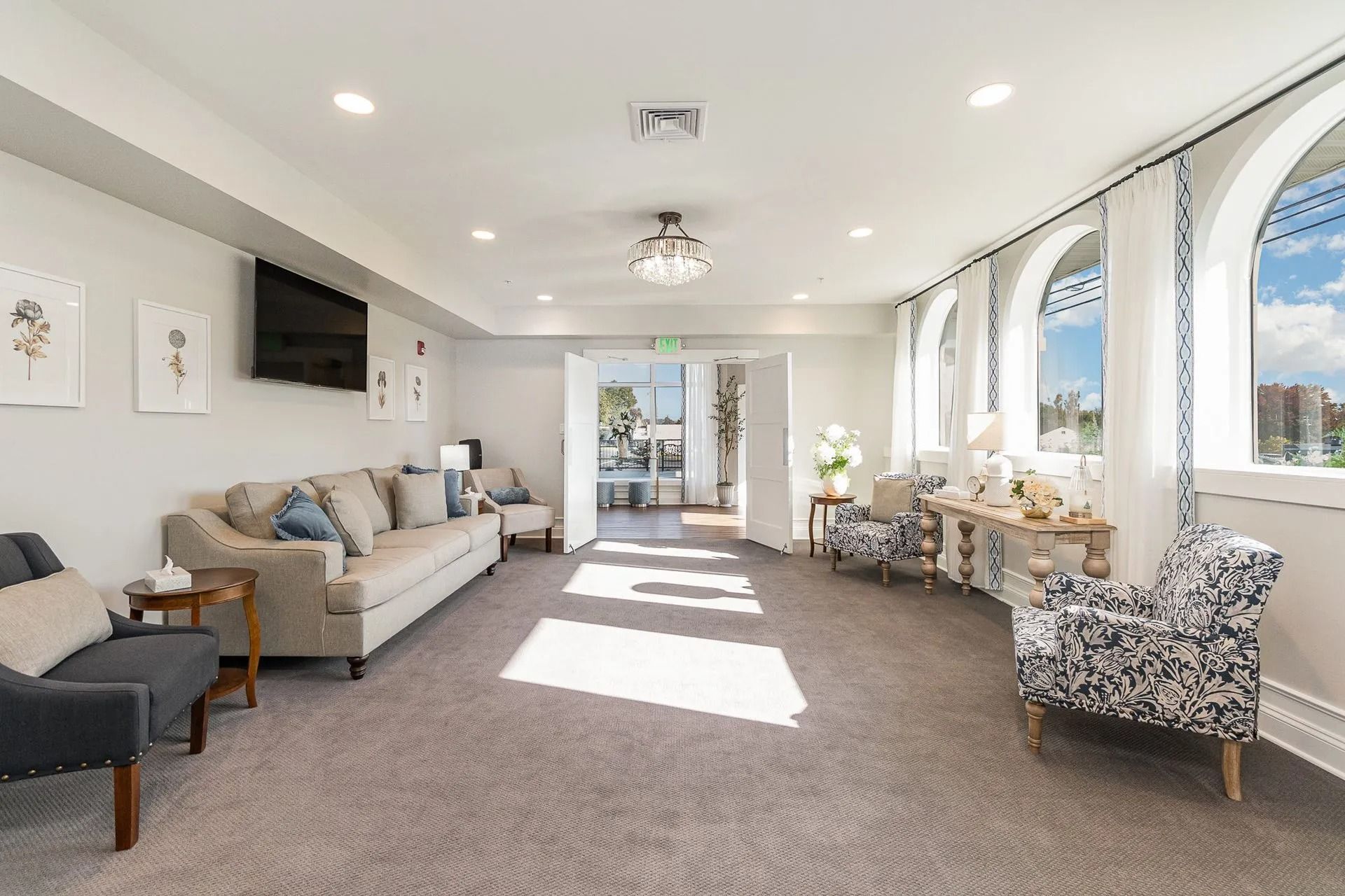 Bright waiting room with arched windows, light-colored furniture, TV, and doors leading outside.