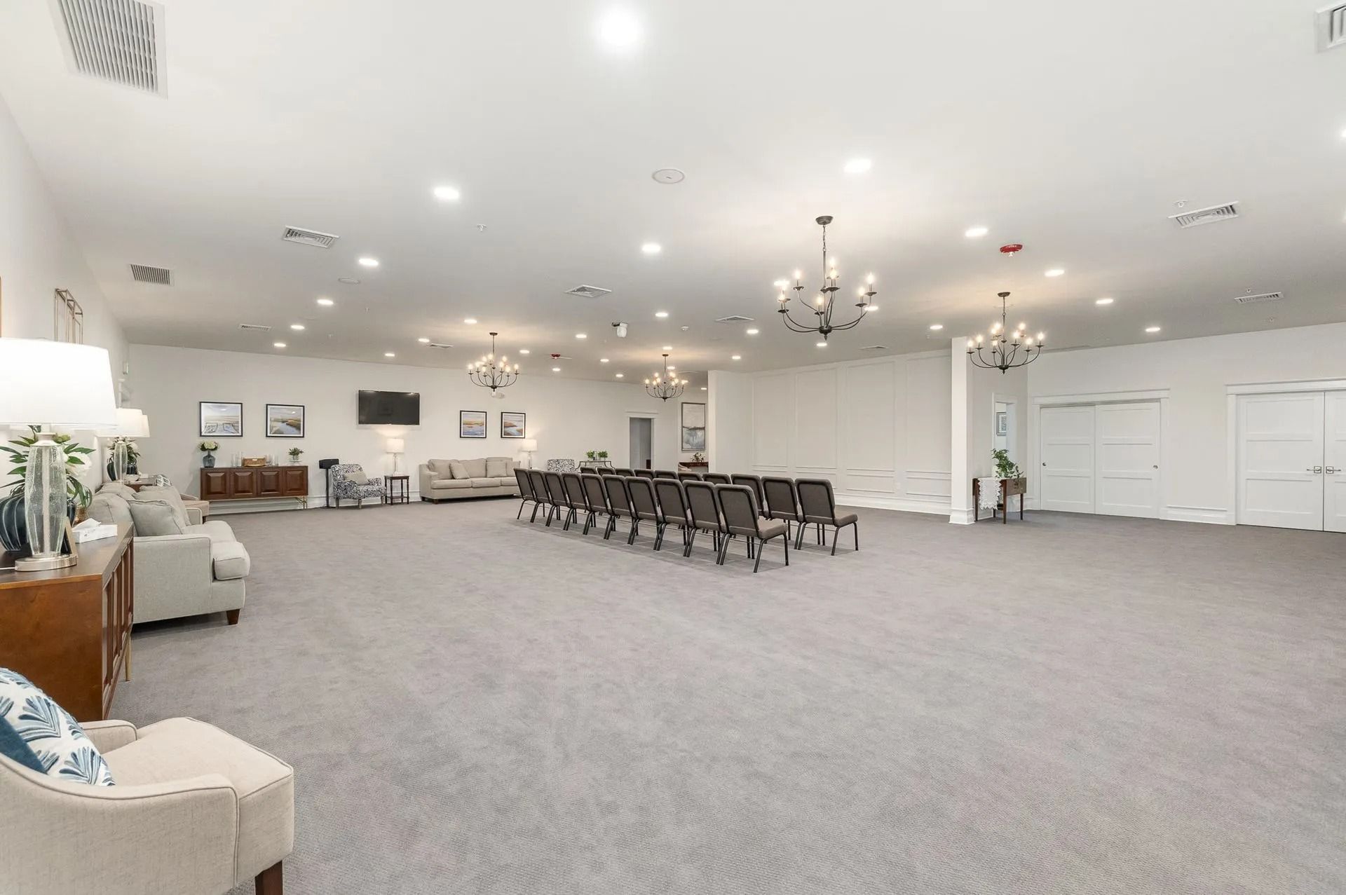 Large, bright room with gray carpet, chandeliers, chairs, and couches; likely a venue for gatherings.