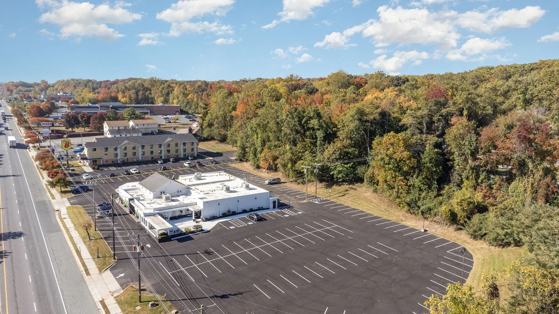 Aerial view of a commercial building with a large parking lot, near a road and a forest with colorful fall foliage.