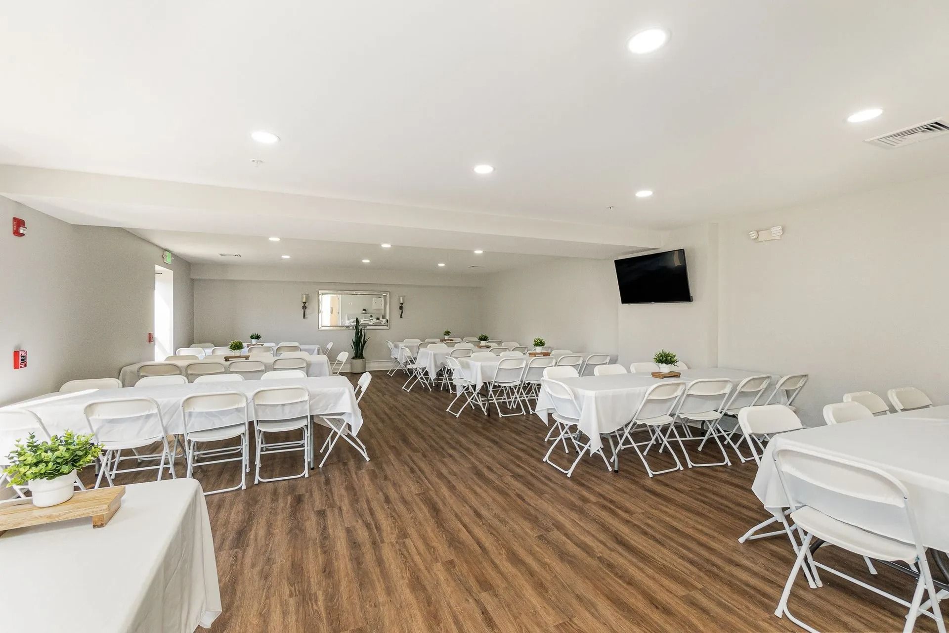 Empty event space with white tables, chairs, and wood flooring; TV on the wall.