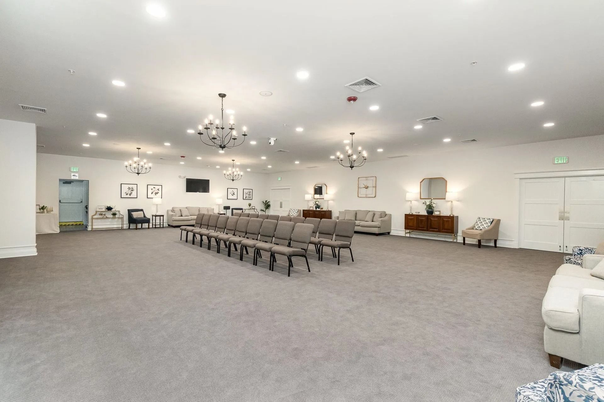 Large, bright room with gray carpet, chairs, sofas, and chandeliers.