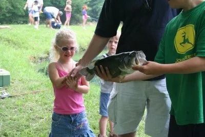Girl reacting to large fish being held by two people near a stream.