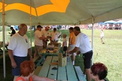People under a tent at an outdoor event, some at a picnic table, interacting with food and drinks.