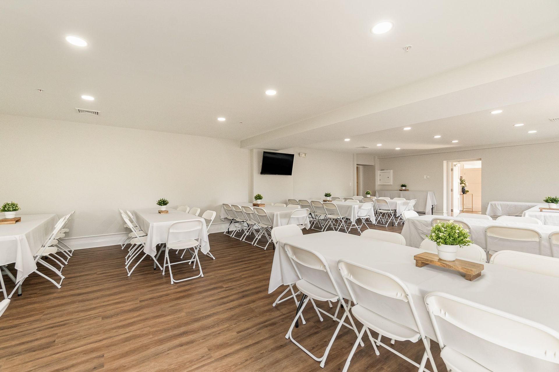 Empty event room with white tables and chairs, wood-look floor, and potted plants.