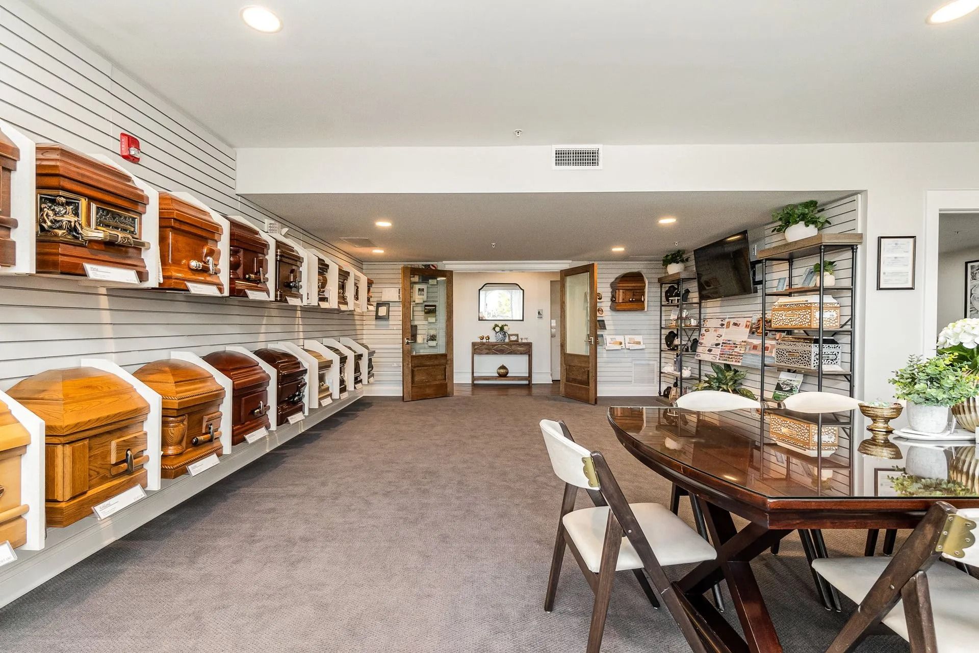 Interior of a funeral home, display of caskets along wall, long wooden table with chairs, carpeted floor.