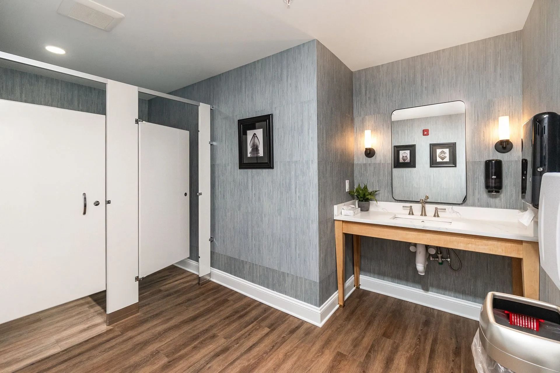 Bathroom with two stalls, a sink, mirror, and paper towel dispenser. Gray textured walls and wooden floor.