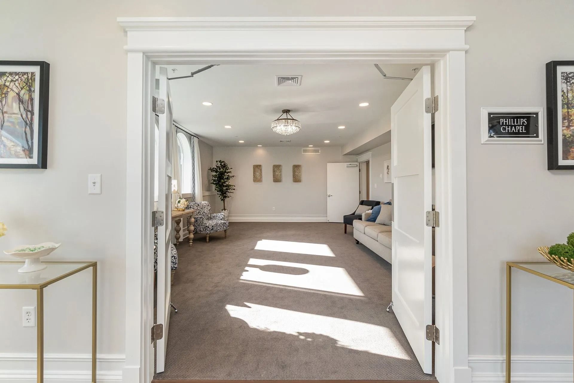 View through doorway into a light-filled room with seating, a chandelier, and framed art.