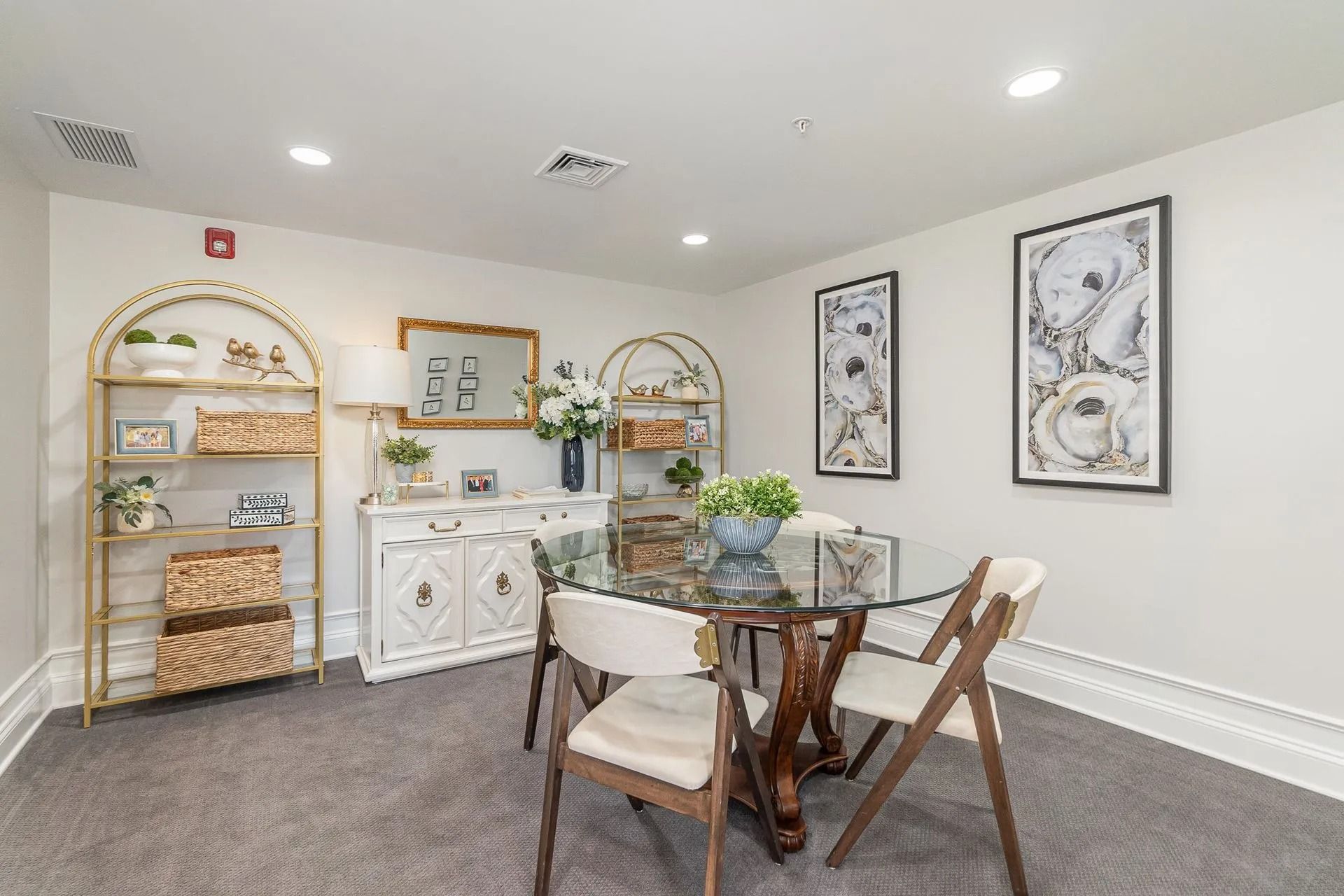 A bright room with a glass-topped table and chairs, a white cabinet, and gold shelving units, with framed art.