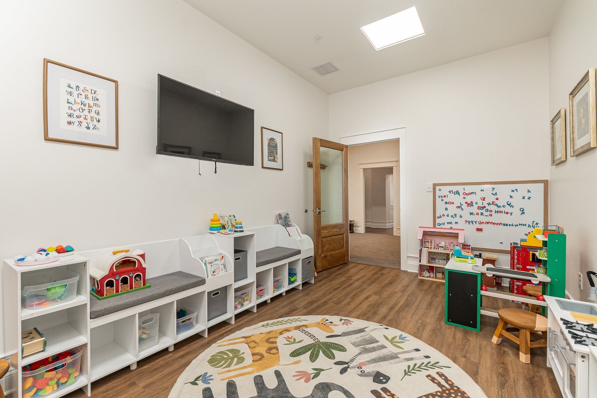 Playroom with toys, benches, TV, and open doorway to another room. A round rug sits in the middle of the floor.