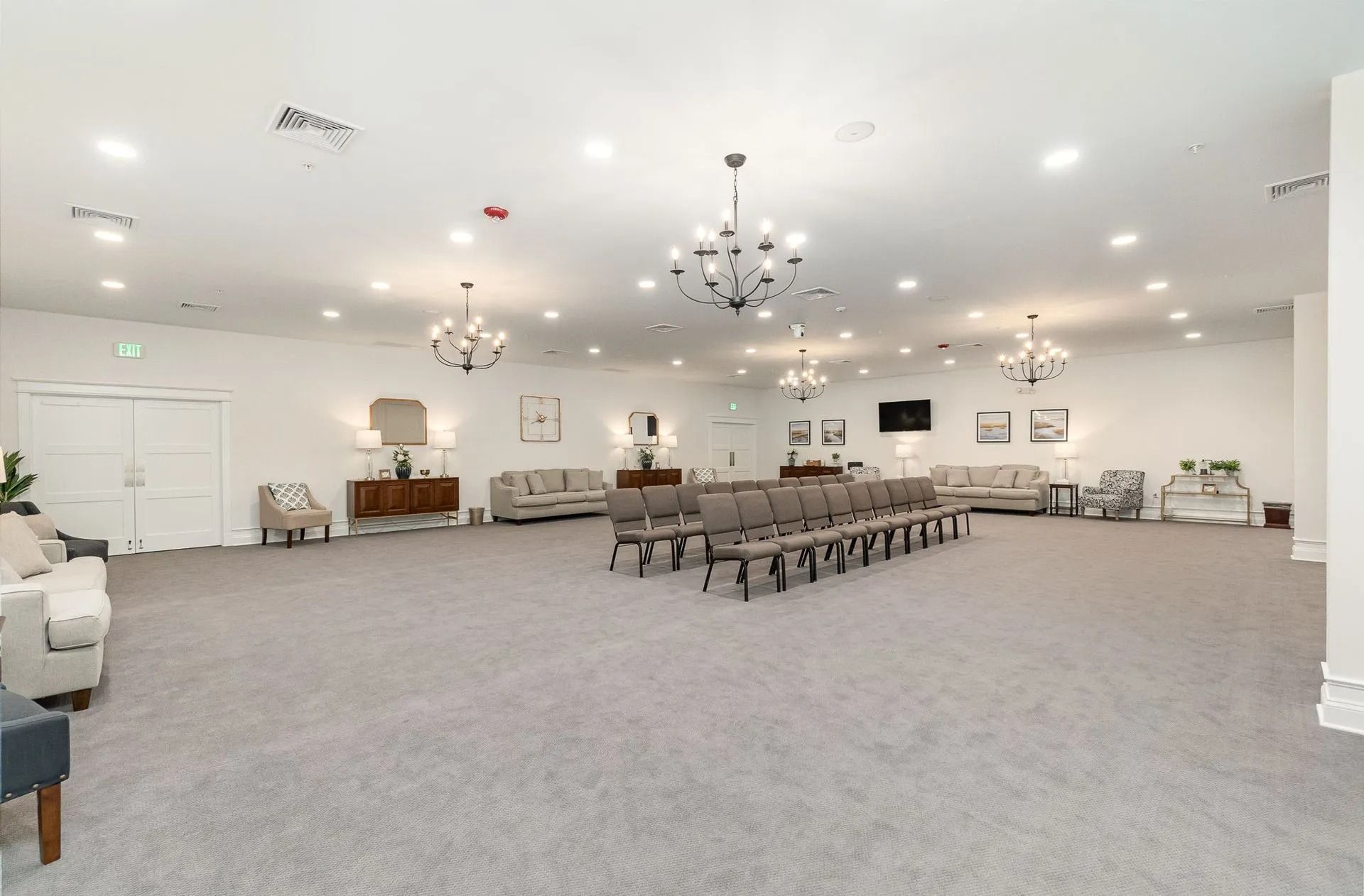Large, well-lit room with gray carpet, sofas, chairs, chandeliers. Likely a funeral home or event space.