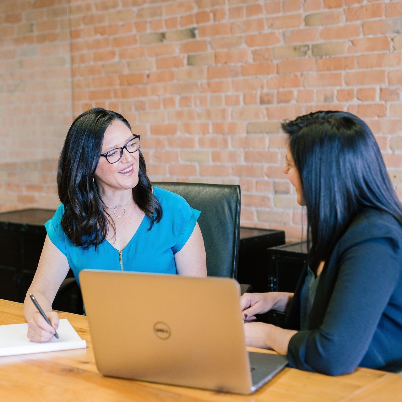 Two women at a table in an office, one writing, the other using a laptop; brick wall in background.