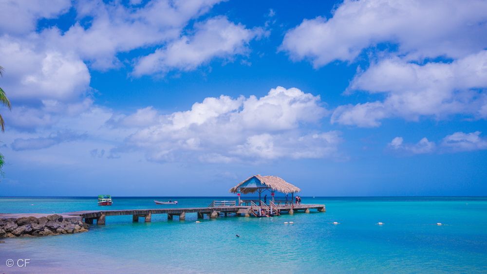 Tropical beach with a wooden pier, a small hut, and turquoise water under a bright blue sky with clouds