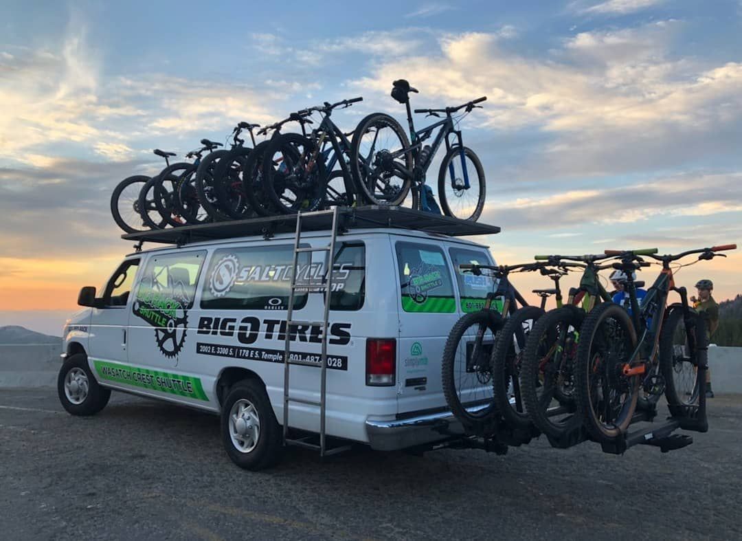 White van loaded with mountain bikes on roof rack and hitch, parked outdoors at sunset.