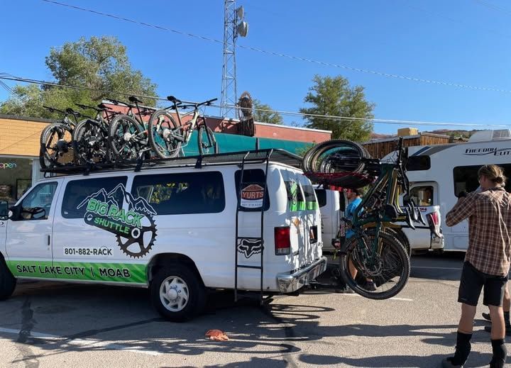 White van with bikes on roof rack, being loaded. Man holding a bike. Daytime outdoors.