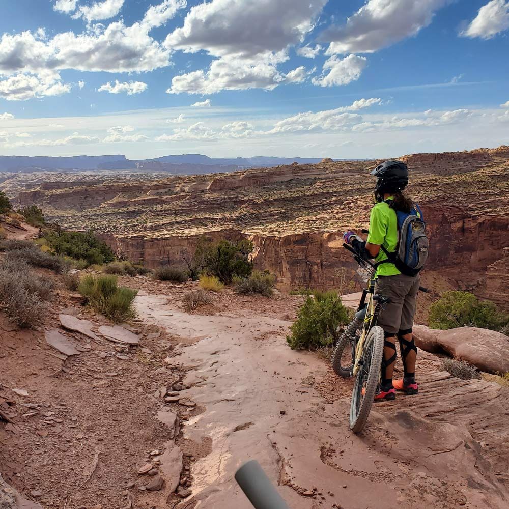 Mountain biker on a red rock trail, overlooking a canyon under a blue sky with clouds.
