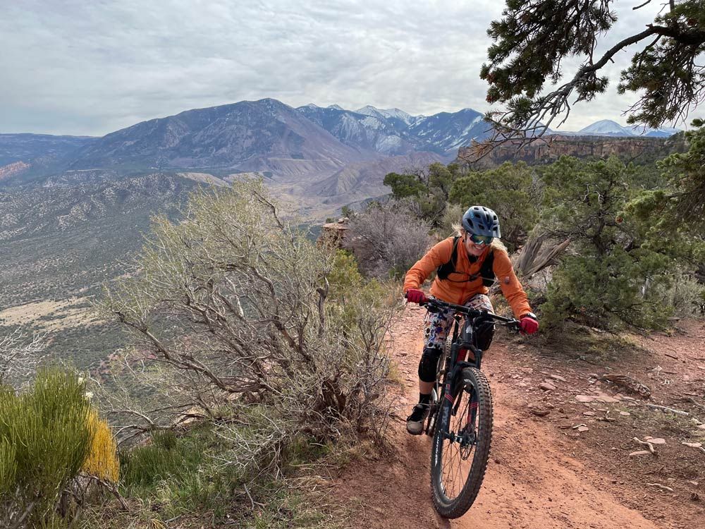 Mountain biker rides red dirt trail with scenic mountain backdrop.