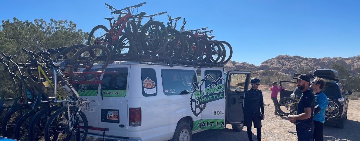 White van loaded with bicycles, people standing nearby in a desert setting.