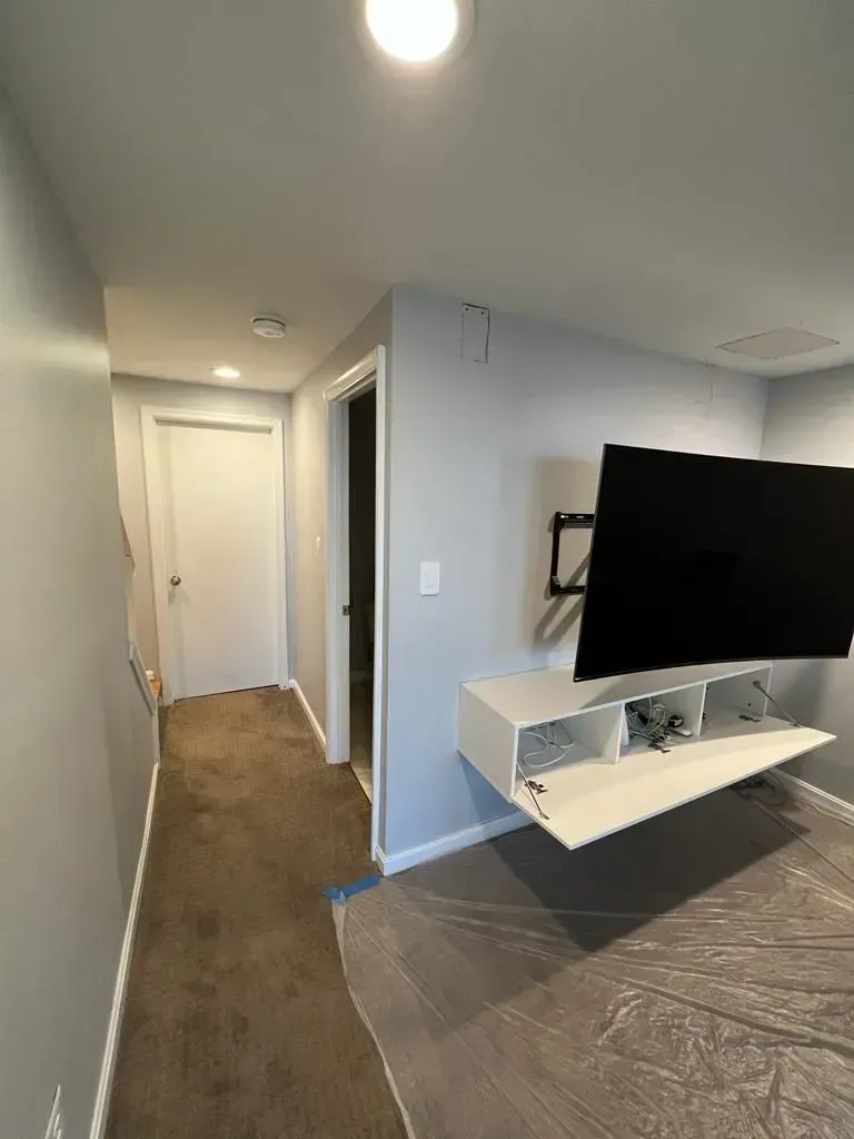 Hallway leading to a door, with wall-mounted TV and white shelf on the right, carpeted floor and covered furniture.