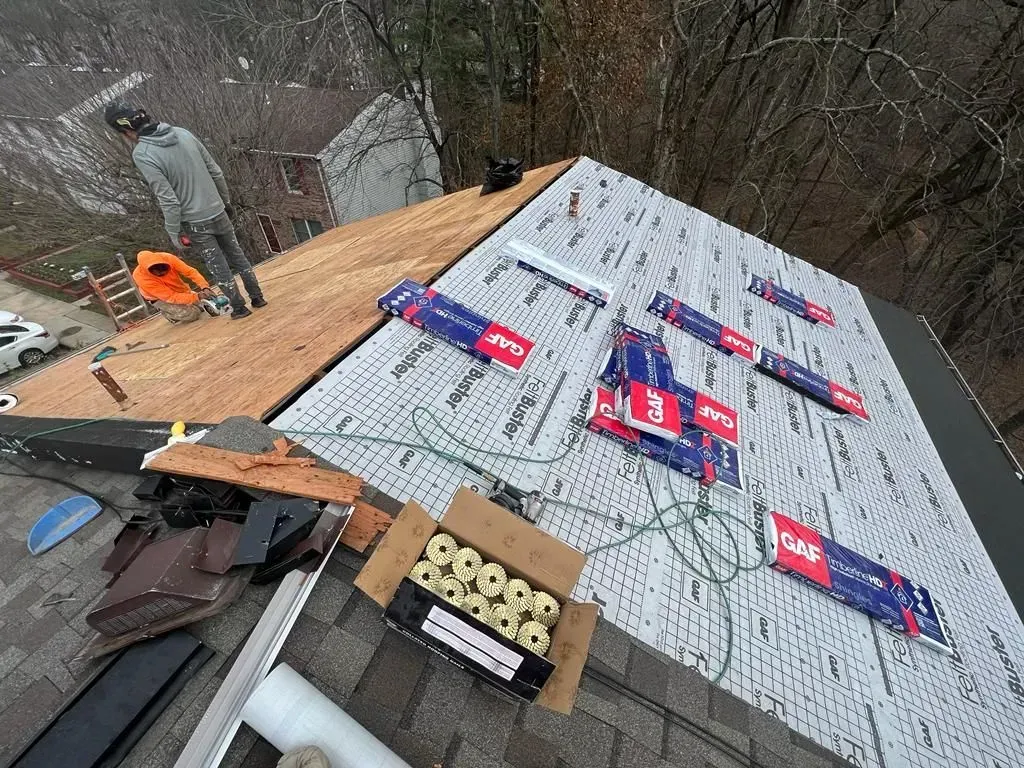 Roof under repair with shingles and roofing materials; worker stands on the left side.
