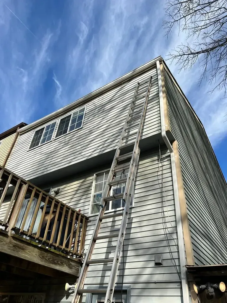 Weathered gray house siding with a wooden deck and ladder under a blue sky