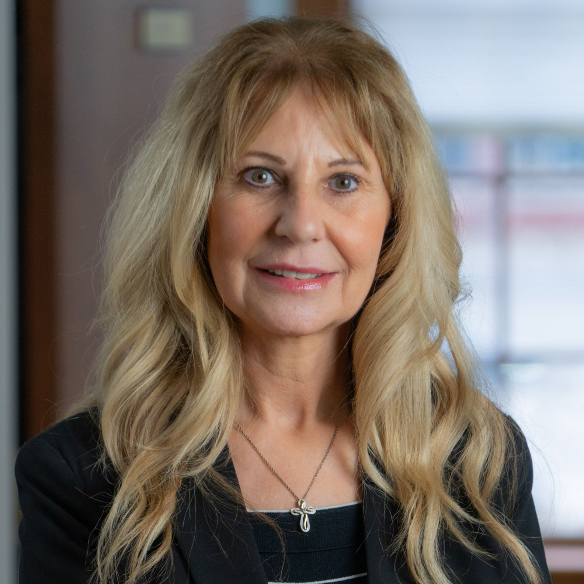 Woman with blonde hair, wearing a black blazer and necklace, smiling in an office setting.