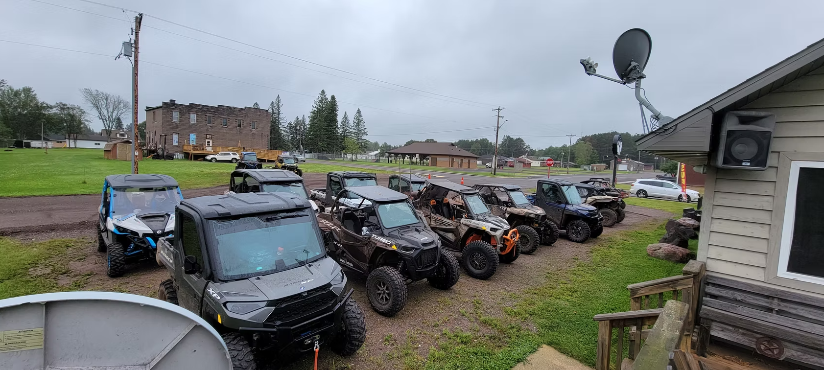 Row of parked off-road vehicles in a muddy lot beside a building under a cloudy sky