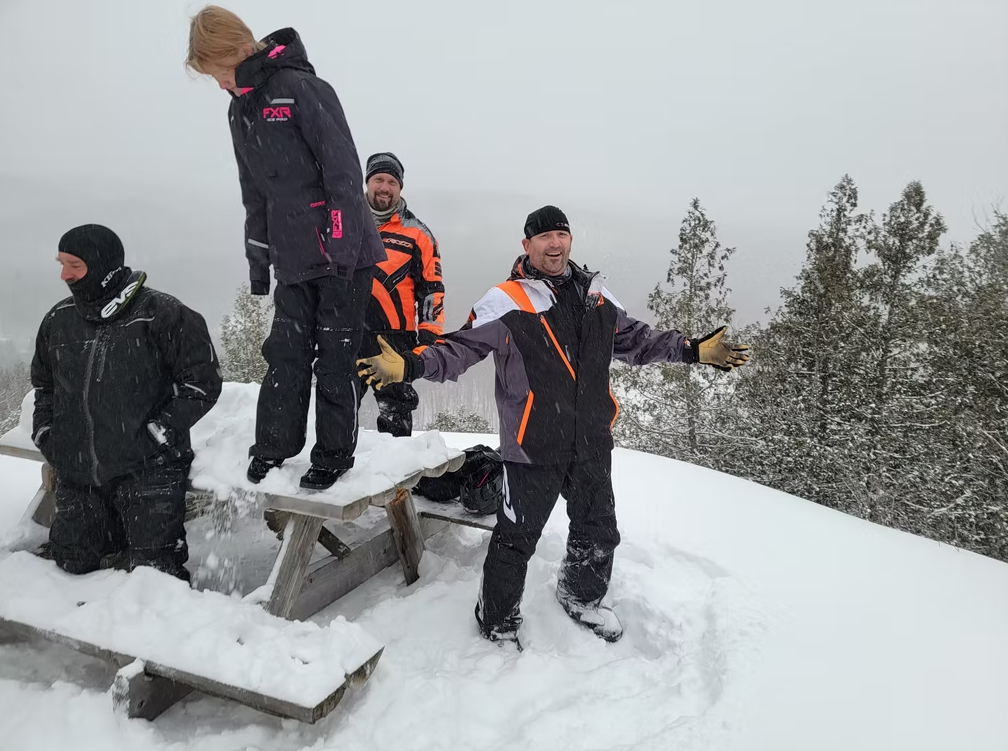 Four people in winter gear standing on a snowy hilltop by a bench, with trees in the background.