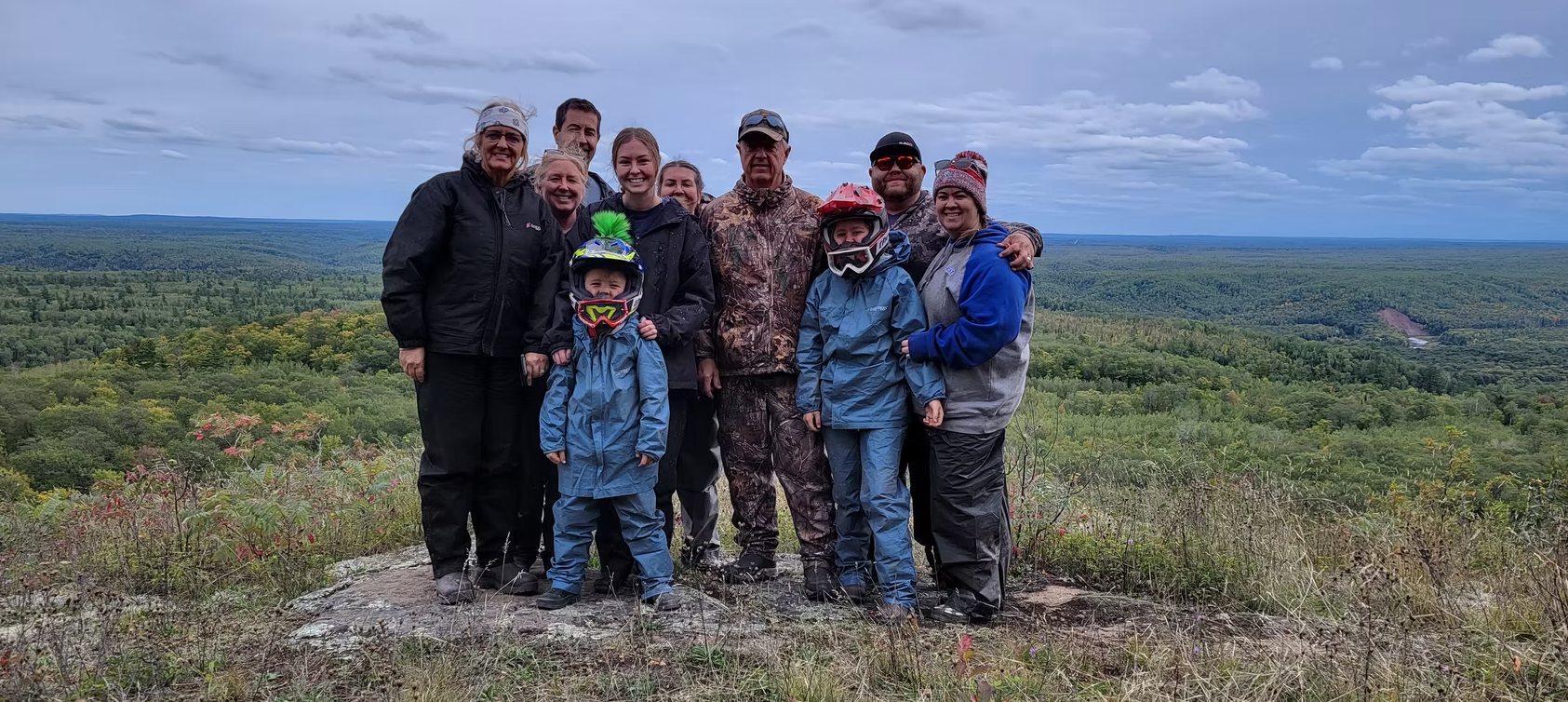 Group of hikers posing on a rocky hilltop with wide forested landscape under a cloudy sky.