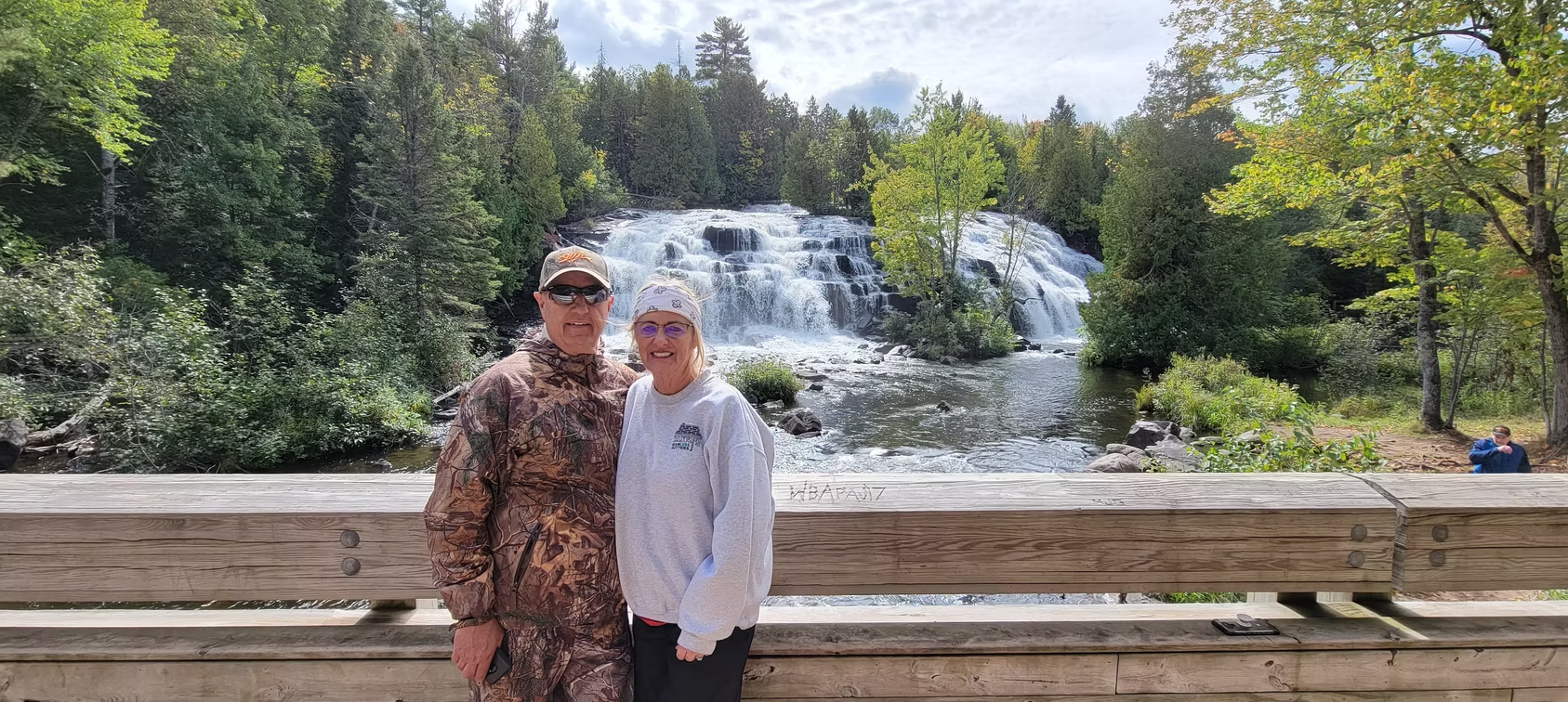 Two people standing by a wooden railing in front of a waterfall in a forested park