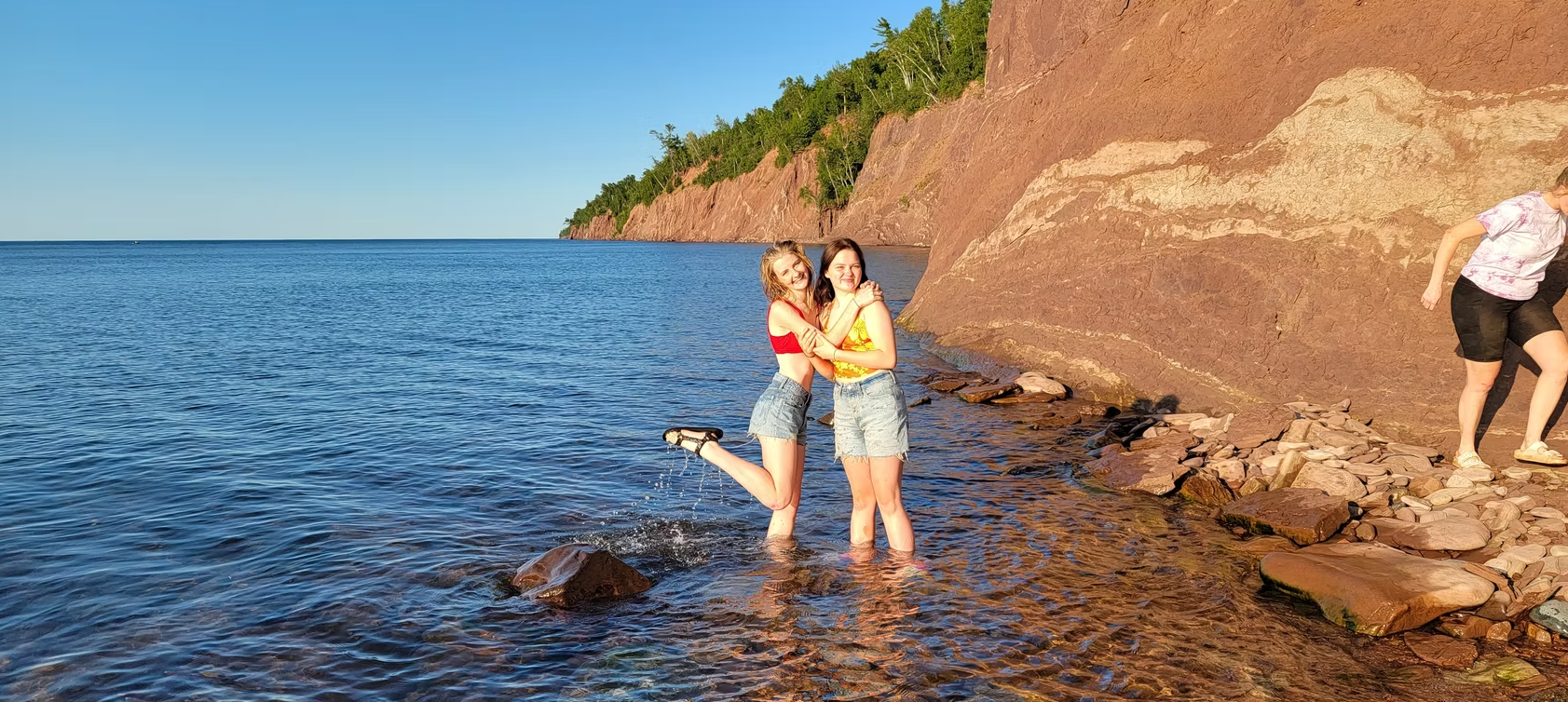 Two people stand in shallow water by a rocky shoreline under a clear blue sky.