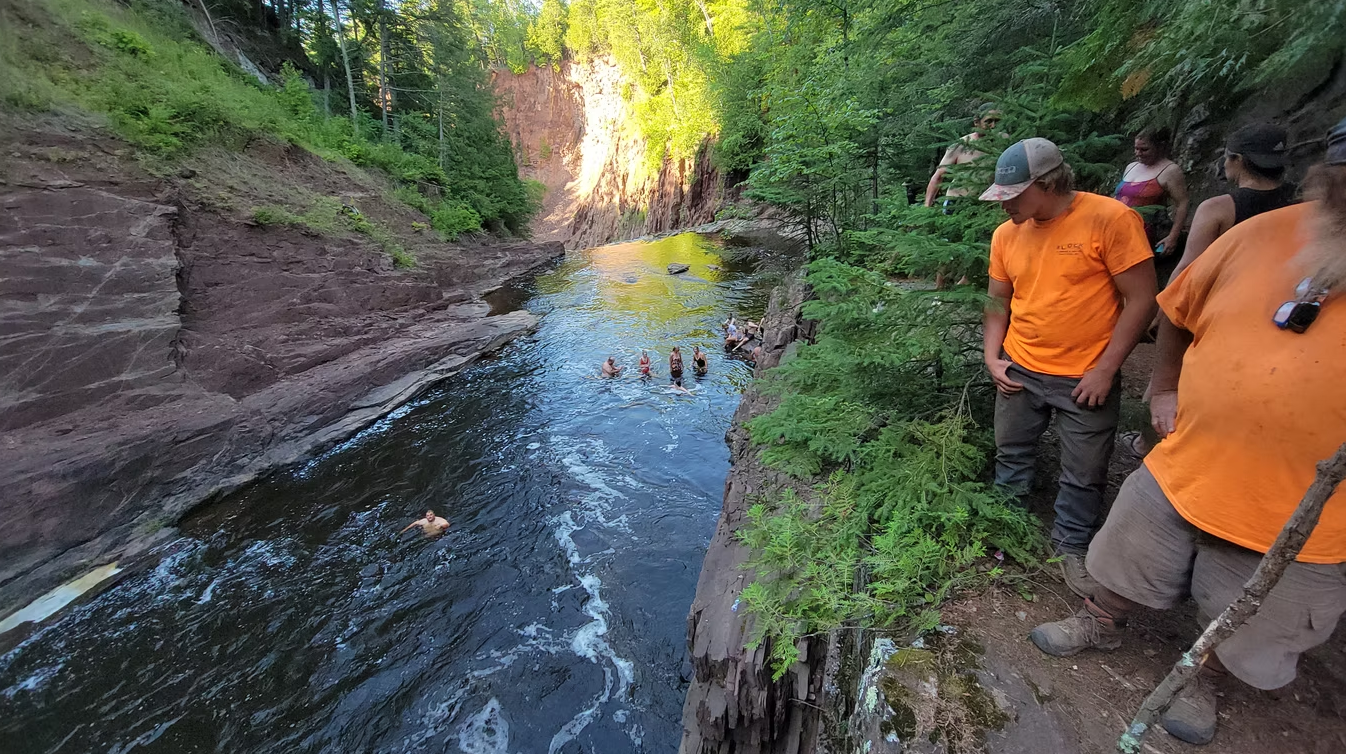 Two hikers in orange shirts stand beside a narrow stream in a lush forest ravine.