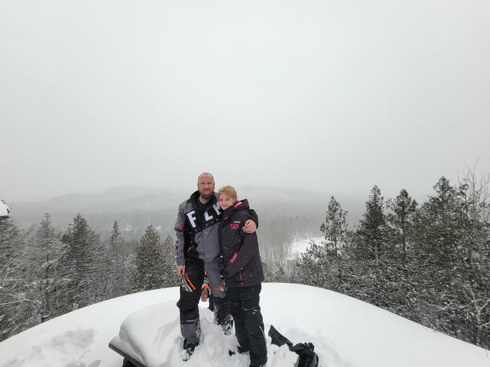 Two people standing on a snowy mountain overlook with snow-covered trees and a foggy sky behind them