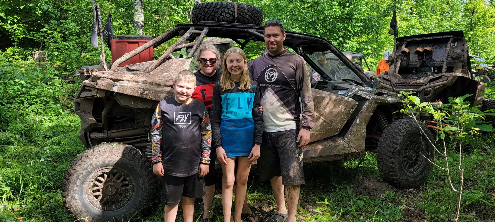 Three people posing beside a muddy off-road vehicle in a green wooded area